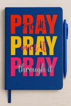 Colorful journal with bold text reading 'Pray on it, Pray over it, Pray through it' in red, yellow, pink, and aqua on a blue cover, with matching blue pen."