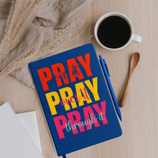 Notebook with 'PRAY on it, PRAY over it, PRAY through it' text on a wooden surface with a cup of coffee and pen.