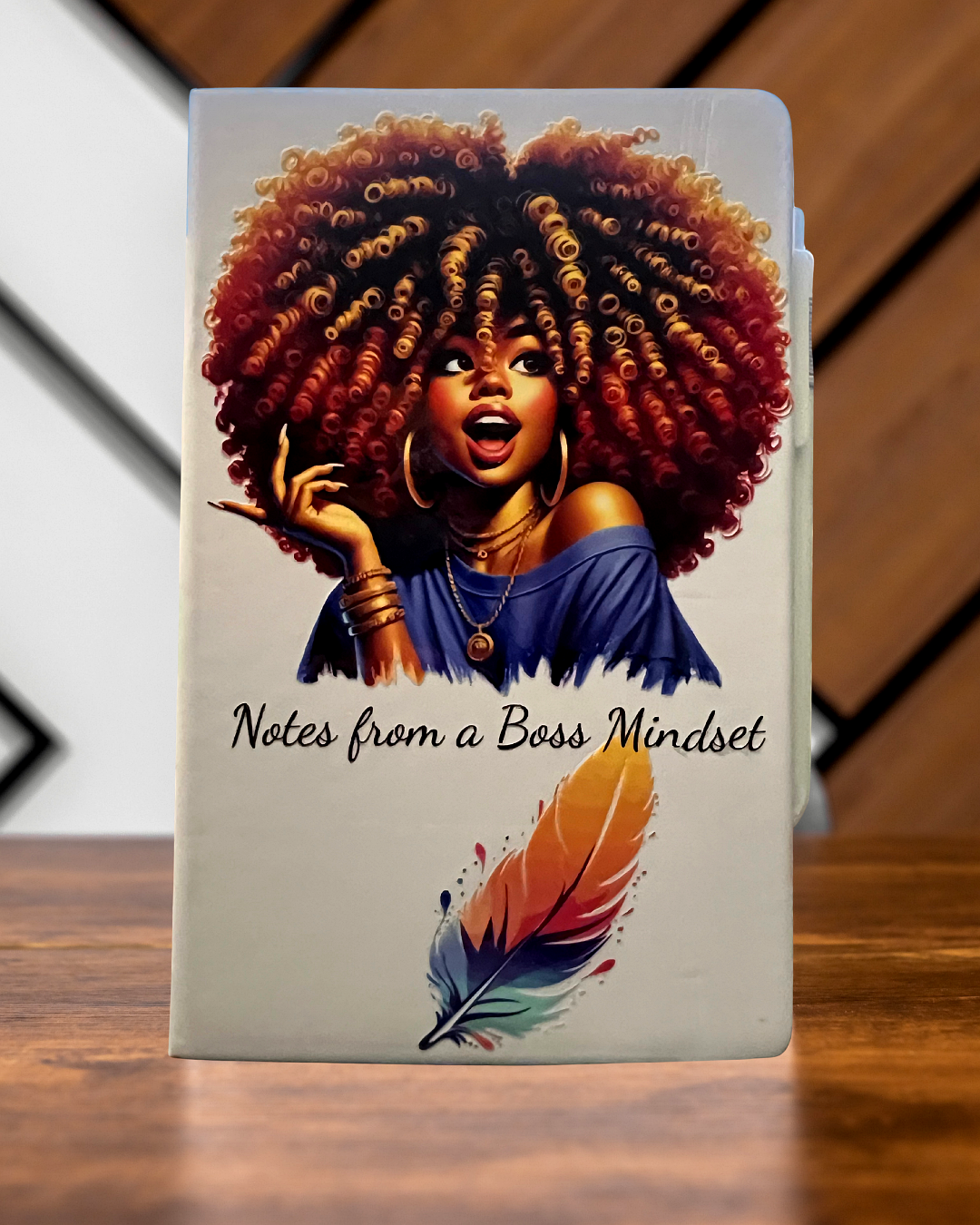 Light blue journal with artwork of a confident Black woman in a blue off-shoulder top, gold jewelry, and voluminous curls, with the words 'Notes from a Boss Mindset' and a colorful feather pen graphic.”
